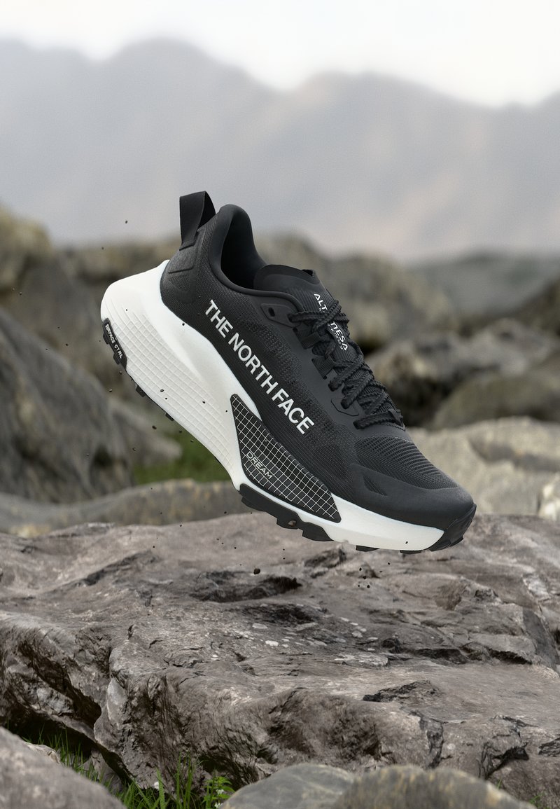 Ασπρόμαυρο παπούτσι τρεξίματος μονοπατιού The North Face που αιωρείται πάνω από βραχώδες έδαφος με θολό ορεινό φόντο.