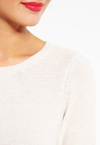 Unterkiefer und Hals einer Frau mit leuchtend rotem Lippenstift, die einen weiß strukturierten Strickpullover vor einem weißen Hintergrund trägt.