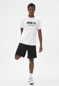 Hvid atletisk t-shirt med "BROKEN NO PAIN NO GAIN" trykt i sort og sorte shorts. Modellen har lyse atletiske sko på.