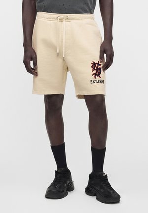 Homme portant un short crème avec un motif brodé et le texte "EST. 1998" sur la jambe gauche, assorti de chaussettes noires et de baskets noires.