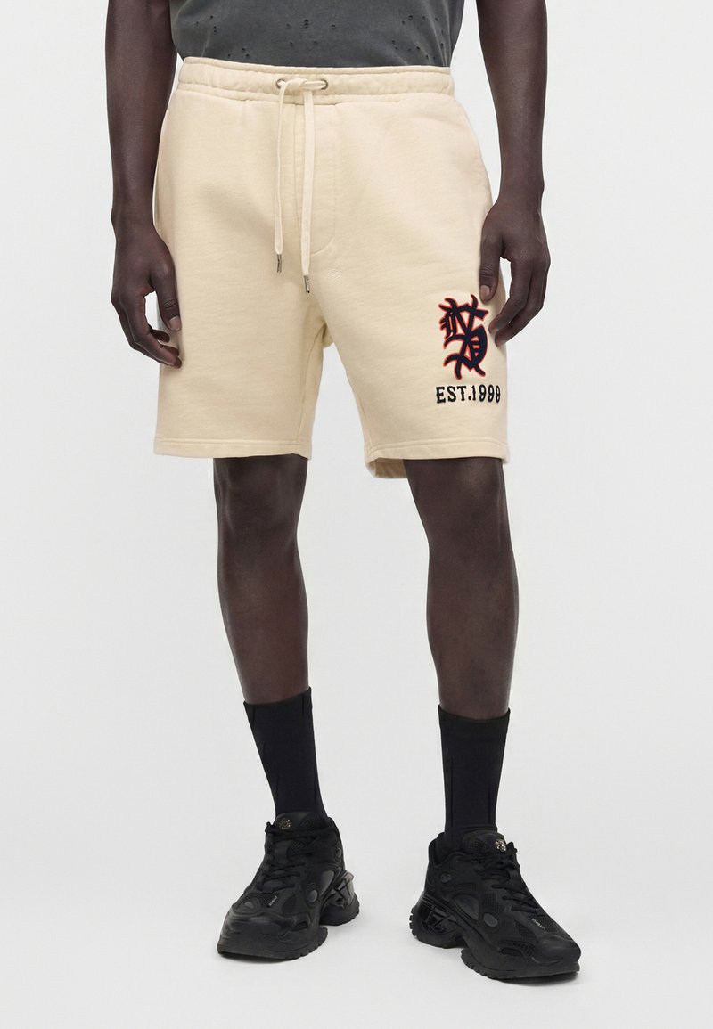 Homme portant un short crème avec un motif brodé et le texte "EST. 1998" sur la jambe gauche, assorti de chaussettes noires et de baskets noires.