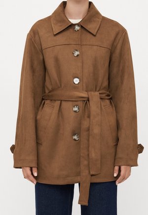 Persona con un abrigo de ante marrón con cinturón, cuatro botones grandes, dos bolsillos delanteros y mangas largas, sobre vaqueros azul oscuro.