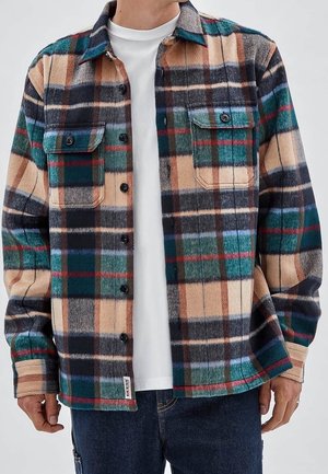 Mężczyzna ubrany w zapinaną na guziki wielokolorową kratowaną flanelową koszulę na białej koszulce i niebieskich dżinsach, stoi z luźno ułożonymi ramionami przy bokach.