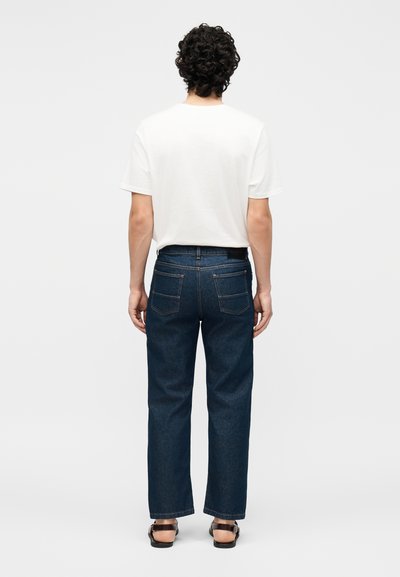 Άνδρας με σγουρά σκουρόχρομα μαλλιά φορώντας ένα λευκό κοντομάνικο μπλουζάκι, σκούρα μπλε τζιν και μαύρα σανδάλια, στέκεται με την πλάτη σε απλό φόντο.