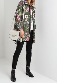 Veste vert olive à motif floral avec une fermeture éclair, associée à un jean skinny noir et des bottines. Accessoirisée d'un sac à main gris clair.