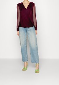 Blusa avvolgente bordò con maniche lunghe trasparenti, abbinata a jeans a gamba larga blu chiaro e tacchi verdi con cinturini. Design semplice, look casual.