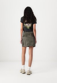 Gonna mini in velluto a coste verde oliva con tasche frontali e posteriori, abbinata a una t-shirt grafica nera e sneakers beige.