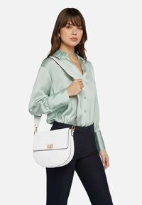 Bolso cruzado de cuero blanco con herrajes dorados, forma curva y tapa plana. El modelo lleva una blusa de satén verde y pantalones azul marino.