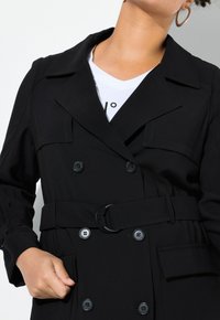 Schwarzer doppelter Trenchcoat mit tailliertem Gürtel, großen Knöpfen und seitlichen Taschen. Mit einem Reverskragen und glatter Stofftextur.