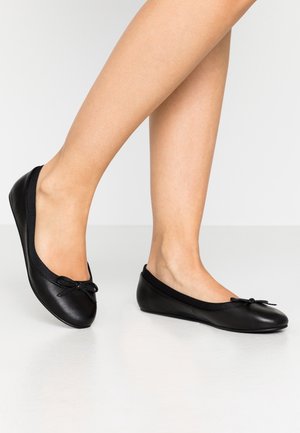 Person trägt schwarze Leder-Ballerinas mit kleinem Schleifendetail, steht auf weißem Boden vor einfarbigem Hintergrund.