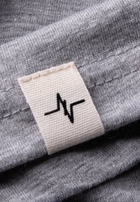 Grauer Stoff mit weicher Textur, versehen mit einem gewebten Etikett, das eine Herzschlaglinie in Schwarz auf einem hellbeigen Hintergrund zeigt.