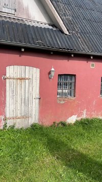 Rdeča zgradba z obrabljenimi belimi lesenimi vrati, z zaklenjenim oknom, zunanjim svetilkom in valovito streho nad zeleno travo.