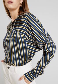 Blouse à manches longues rayée bleu marine et jaune avec un col rond, tissu texturé et détails froncés aux épaules. Pantalon blanc porté.