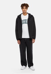 Svart zip-up jacka med huva, tillsammans med en vit t-shirt med turkosa texter, och lösa svarta jeans. Vita sneakers fulländar outfiten.