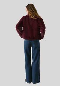 Jachetă din blană sintetică burgundy cu guler înalt și mâneci lungi, asortată cu blugi blue denim cu picioare largi, văzută din spate.