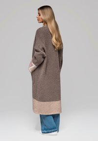 Cardigan marrone a maglia con maniche lunghe, caratterizzato da un motivo pastello ai polsini e un orlo inferiore beige. Abbinato a jeans di denim blu.