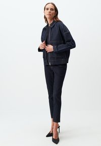 Giacca in denim con zip frontale, tasche applicate e vestibilità ampia di colore blu scuro. Abbinata a jeans skinny scuri e scarpe con tacco alto nere.