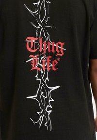 Čierne bavlnené tričko s červeným textom "Thug Life" a bielym grafickým dizajnom pripomínajúcim tŕne na chrbte. Krátke rukávy, okrúhly výstrih.