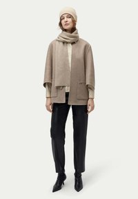 Beige ullblandad capejacka med fickor, matchad med en halsduk och en beige stickad mössa. Svarta läderbyxor och spetsiga svarta stövlar fulländar uppsättningen.