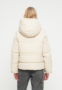 Puffervest i en lys beige farve med en blød, mat finish. Har en stor, påsat hætte og vandret quilted detaljer.