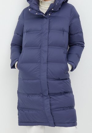 Kobieta ubrana w długi, granatowy pikowany płaszcz z bocznymi kieszeniami i kapturem, na białym golfie.