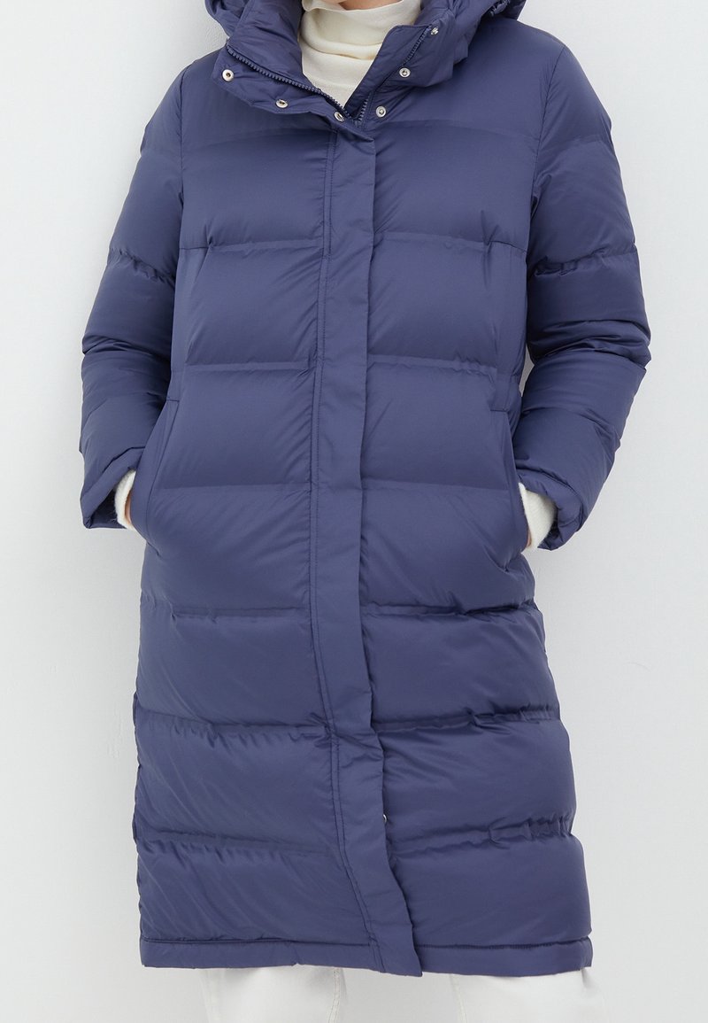 Mujer con un abrigo acolchado largo de color azul marino, con bolsillos laterales y capucha, sobre un suéter blanco de cuello alto.