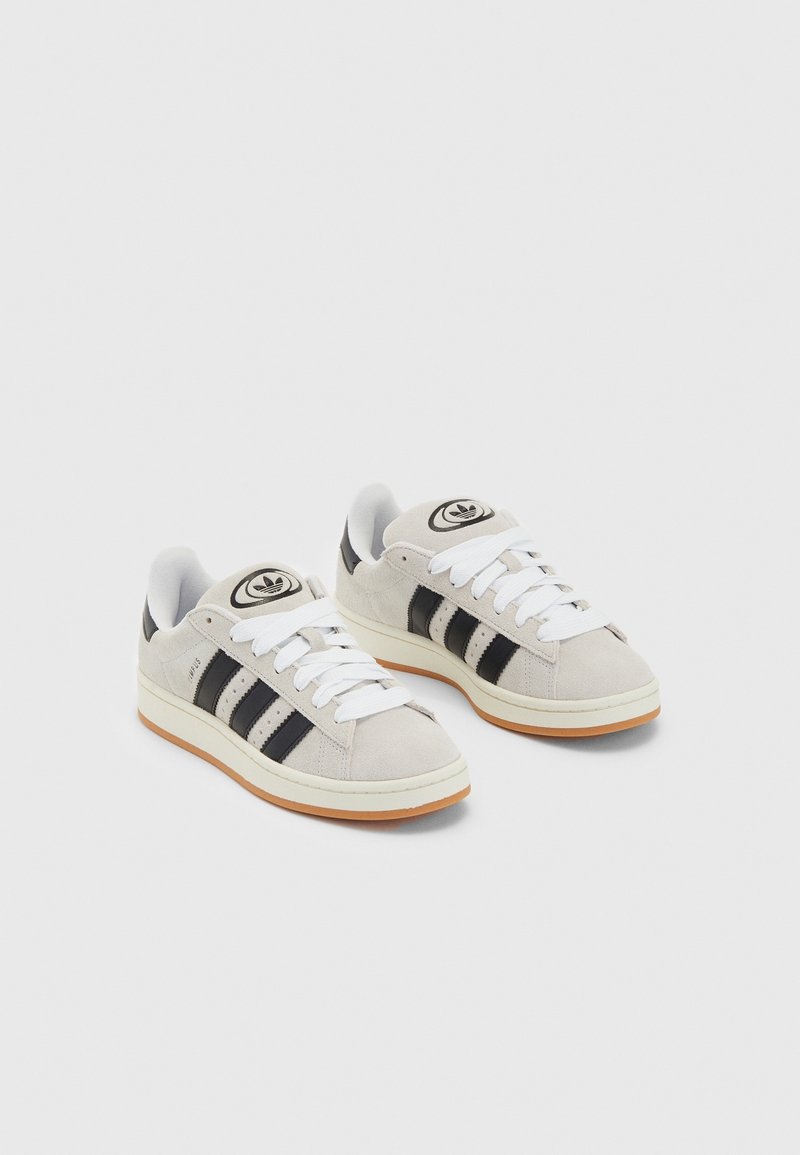 Une paire de sneakers en suède gris clair avec des rayures noires, des lacets blancs, des semelles de couleur gomme et le logo Adidas sur les languettes, sur fond blanc.