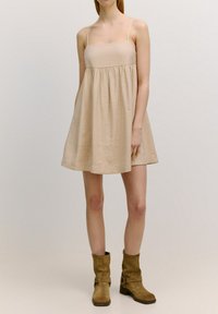 Beige mouwloos jurkje met een vierkante halslijn, bijeen geraapt taille en geflonkete rok. Draag met bruine laarzen tot de knie. Glad, lichtgewicht stof.