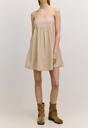 Beige mouwloos jurkje met een vierkante halslijn, bijeen geraapt taille en geflonkete rok. Draag met bruine laarzen tot de knie. Glad, lichtgewicht stof.