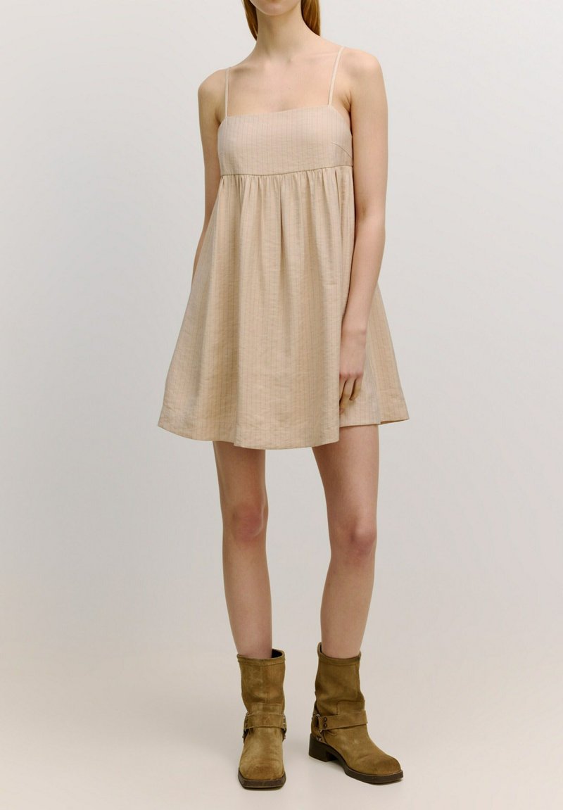 Beige mouwloos jurkje met een vierkante halslijn, bijeen geraapt taille en geflonkete rok. Draag met bruine laarzen tot de knie. Glad, lichtgewicht stof.