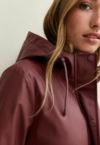 Vinröd vattentät jacka med huva, med en texturerad yta, dragsko och tryckknappar på kragen för säker stängning.