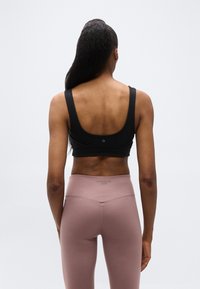 Γυναίκα με μακριά μαλλιά που στέκεται  με την πλάτη προς τα εμπρός, φοράει μαύρο αθλητικό σουτιέν και μωβ κολάν ψηλής μέσης σε ουδέτερο φόντο.