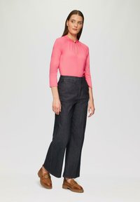 Vaaleanpunainen istuva toppi, jossa on avaimenreikäyksityiskohta, yhdistettynä tummiin denim-korkeavyötäröisiin leveälahkeisiin housuihin ja ruskeisiin loafer-kengiin, joissa on metallikoristeluja.