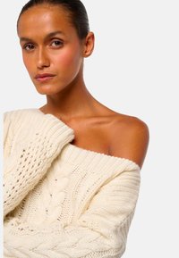 Frau mit glatter, dunkler Haut, die einen cremefarbenen, schulterfreien Zopfmuster-Pullover trägt, vor einem schlichten weißen Hintergrund.