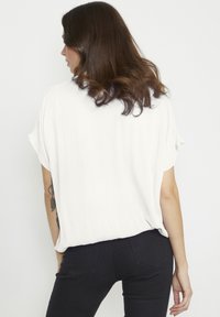White short-sleeve blouse with a crepe texture, featuring a loose fit and a rounded hem, paired with fitted black jeans.
