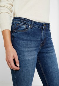 Tummansiniset farkut, joissa on kapea malli, etutasku, ommeltu yksityiskohtia sekä klassinen nappi- ja vetoketjusulku.