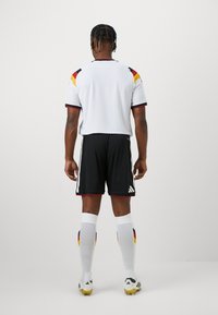 Σέτ ποδοσφαίρου Adidas με λευκή μπλούζα κοντά μανίκια, μαύρες βερμούδες και λευκές κάλτσες με κόκκινες, κίτρινες και μαύρες πινελιές.