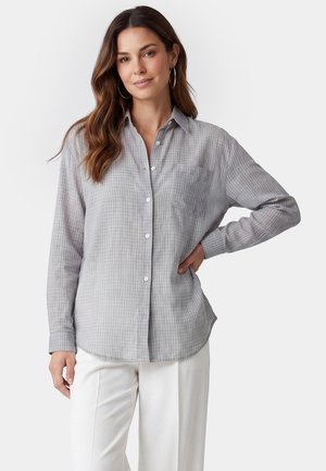 Vrouw met lang bruin haar die een grijs geruite blouse met knopen en een witte broek draagt, poserend met één hand op haar heup tegen een witte achtergrond.