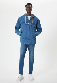 Chaqueta de mezclilla con capucha, cordones naranjas y texto bordado "¿Y QUÉ?", bolsillo tipo canguro en la parte delantera, combinada con jeans azules ajustados y zapatillas blancas.