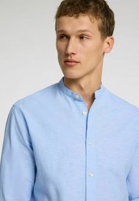 Svetlo modra, dolgimi rokavi majica z ovratnikom, s stiskanjem gumbov in teksturirano tkanino. Standardna kroja z minimalnimi detajli.