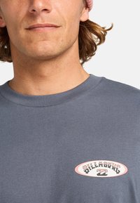 Camiseta gris de manga corta con cuello redondo y cuello acanalado, que presenta un colorido logotipo gráfico en el pecho. Textura de tela de algodón suave.