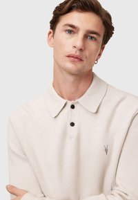 Pull beige clair en tricot avec un col à revers, trois boutons noirs et un petit logo gris. Texture douce et coupe décontractée.