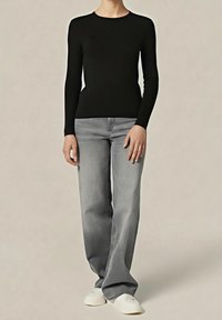 Personne portant un haut noir à manches longues, un jean gris à jambes larges et des chaussures blanches, debout devant un fond uni.