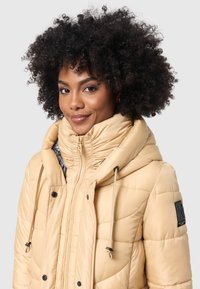Puffy beige jacket with a high collar, zip closure, and drawstrings. Features black buttons and a logo patch on the sleeve, smooth texture.