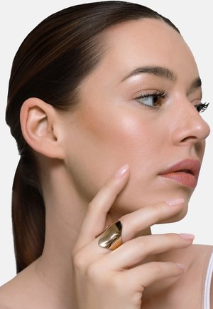 Donna con capelli scuri raccolti dietro, che si tocca il viso con le dita, indossa un grande anello d'oro lucido al dito medio, guarda verso destra.