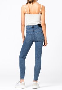 Mujer vestida con jeans ajustados de tiro alto en azul, top blanco de tirantes y zapatillas blancas, de pie con la espalda hacia la cámara.