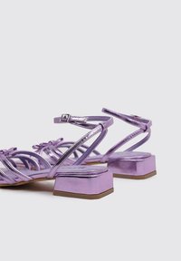 Sandalias de tacón cuadrado metálicas moradas con tiras finas y acentos de lazo. Presentan una suela texturizada y un diseño de punta cuadrada.