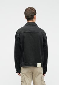Sort denimjakke med spids krave, lange ærmer og sidelommer. Har et hvidt mærke ved bagste kant. Glat overflade.