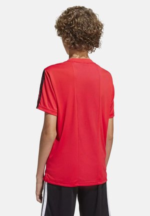 Personne aux cheveux bouclés portant une chemise rouge à manches courtes et un pantalon noir avec des bandes blanches, dos tourné sur un fond uni.