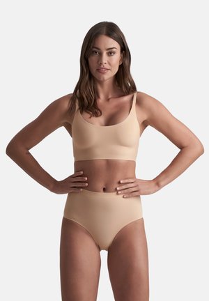 Vrouw met lang bruin haar, gekleed in een naadloze beige beha en hoge tailleonderbroek, staande met de handen op de heupen tegen een effen achtergrond.